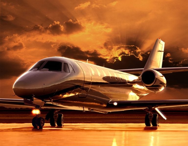 A sleek private jet is parked on an airstrip at sunset, with the setting sun casting a warm, golden glow across the scene. The sky is partly cloudy, allowing beams of sunlight to break through and illuminate the aircraft's shiny exterior.