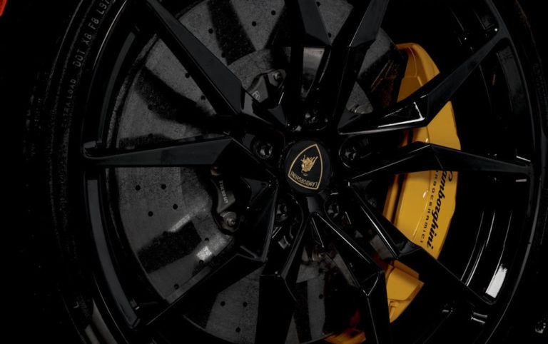 Close-up view of a Lamborghini car wheel, featuring black multi-spoke rims with the Lamborghini logo in the center. Visible behind the wheel is a yellow brake caliper with the Lamborghini name written on it.