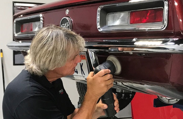 In the Revivify Coatings Gallery, a person is meticulously polishing the rear bumper of a classic maroon car with an electric buffer. The back of the car flaunts an "SS" emblem as they concentrate on their task, with various tools and car products scattered in the background.