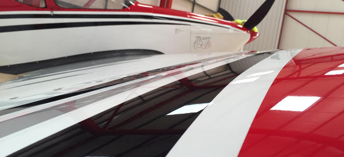 Close-up view of two adjacent small aircraft inside a hangar. The foreground features the wing of a red and white plane with a glossy surface, reflecting the hangar lights. Another plane with a white exterior and black stripes is partially visible behind it.