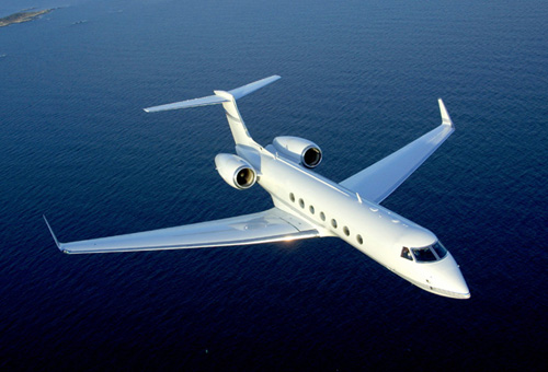 A sleek, white private jet with twin engines flies over a vast body of dark blue water. The aircraft's streamlined design and glossy finish are clearly visible as it glides smoothly, with subtle landmasses faintly visible in the background.