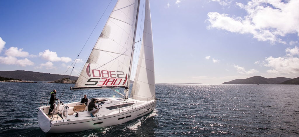 A white sailboat glides on a shimmering body of water under a blue sky with scattered clouds. Two people are on the deck, and the sails are fully raised. Hills and distant landmasses are visible on the horizon. The sailboat's identification number, "10882 S3801," is visible on the sail.