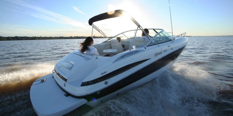 A white motorboat with black trim is speeding on a lake or ocean with two people on board. One person is seated at the back, while the other is seated at the front, facing forward. The sun is low in the sky, creating a bright reflection on the water.