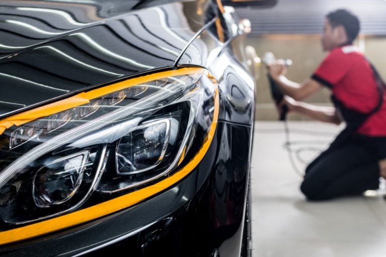 A close-up shot of a black car's headlight being polished, with the hood and front corner in view. In the blurry background, a person in a red shirt kneels while working on the car’s exterior. The lighting reflects off the car’s shiny, polished surface.