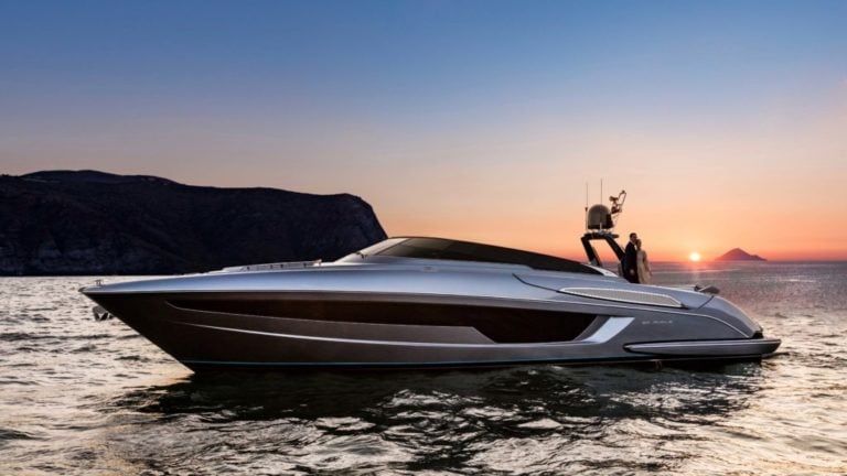 A sleek, modern motor yacht is anchored on calm waters during a sunset. The sun is setting behind a mountainous landscape, casting a warm glow over the scene. The yacht features a streamlined design and reflective surfaces.