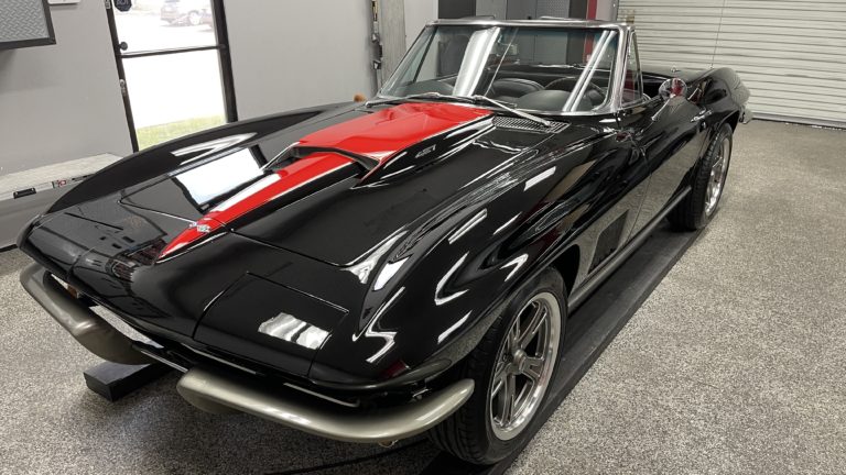 A classic black convertible sports car with a red stripe on the hood is parked indoors. The car features a sleek design with a shiny exterior, and the interior is visible through the open top. The room has a grey and white color scheme with a garage door in the background.