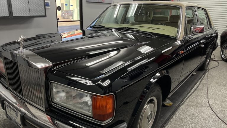 A polished black luxury car with a beige roof, likely a Rolls-Royce, is parked indoors. The car has a distinctive hood ornament, chrome grille, and vintage styling. Nearby, equipment and tools are visible, suggesting the setting is a garage or workshop.