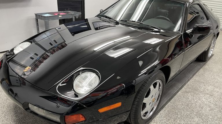 A black Porsche 928 sports car is parked in a garage with a gray speckled floor. The car's sleek, aerodynamic body shines under the overhead lights, and its front left headlight and emblem are prominently visible. The car has polished chrome wheels.