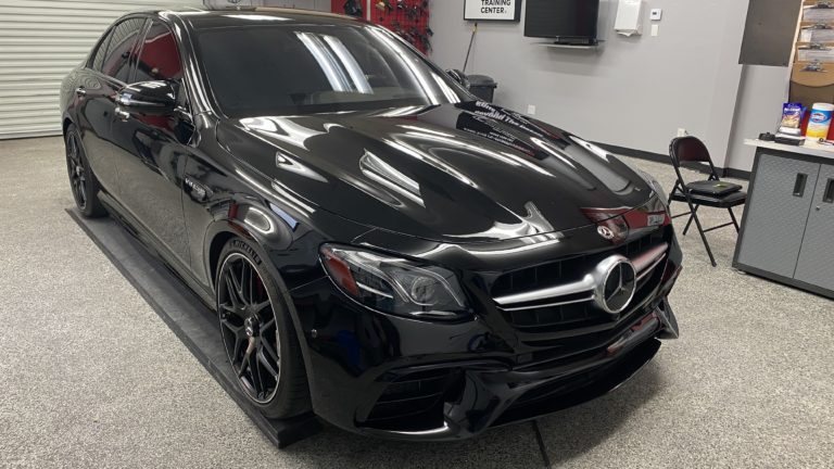 A sleek, black Mercedes-Benz sedan is parked indoors on a garage-like floor. The car features tinted windows, glossy finish, and black alloy wheels. Various tools and equipment are visible on the walls in the background.