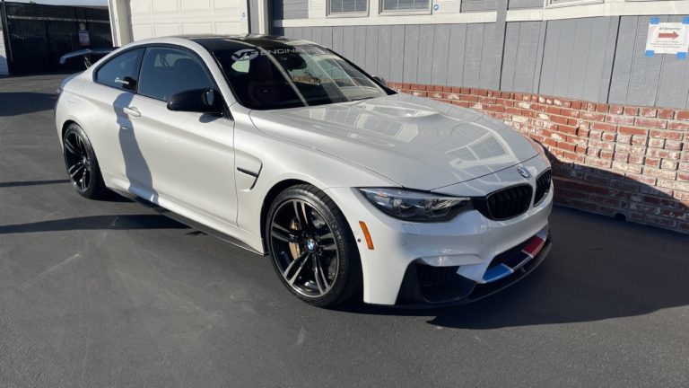 A sleek, white BMW coupe is parked on a smooth, paved surface near a building with a brick and gray exterior. The car features black alloy wheels and colorful decorative stripes on the front bumper. The sun casts soft shadows, highlighting the car's contours.