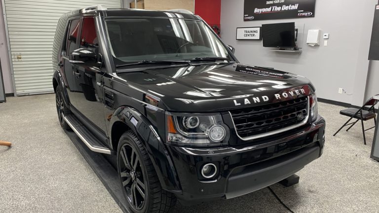 A shiny black Land Rover SUV is parked in a well-lit auto detailing shop. The vehicle is positioned in front of a training area, with various detailing tools and equipment visible around the workspace. The car's exterior appears spotless and polished.