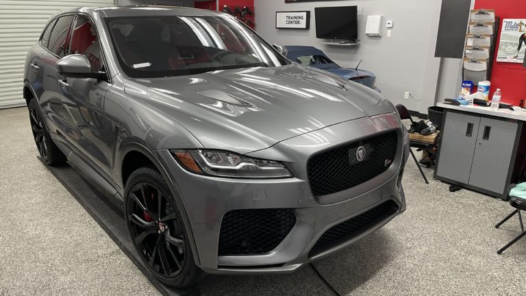 A sleek silver Jaguar SUV is parked inside a garage. The vehicle has a black grille and rims, and the garage contains various tools, equipment, and a "Training Center" sign on the wall. The environment is clean and well-organized.