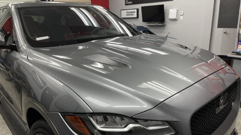 A sleek, silver car is parked in a modern garage or showroom. The vehicle has a black grille with a jaguar emblem, and the hood features two noticeable vents. The environment includes a wall-mounted TV and a "TRAINING CENTER" sign. The area is clean and well-lit.