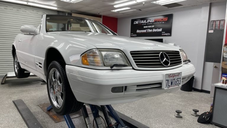 A white convertible Mercedes-Benz with California license plates is elevated on a lift in an auto detailing shop. The sign in the background reads "The Detailing Pros Auto Care Center." Various tools and equipment are visible around the vehicle.