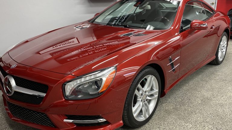 A shiny red Mercedes-Benz sports car is parked in a clean, well-lit garage. The car has a sleek design with aerodynamic curves, silver alloy wheels, and a glossy finish that reflects the light. The Mercedes-Benz logo is prominently displayed on the front grille.