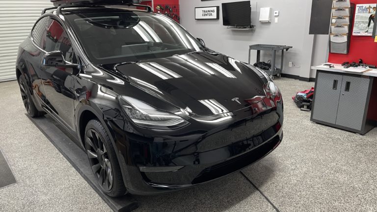 A sleek, black Tesla Model Y parked inside a modern garage or workshop. The car is positioned facing forward with a roof rack installed. The garage has grey floors, red and grey walls, and various tools and equipment in the background.