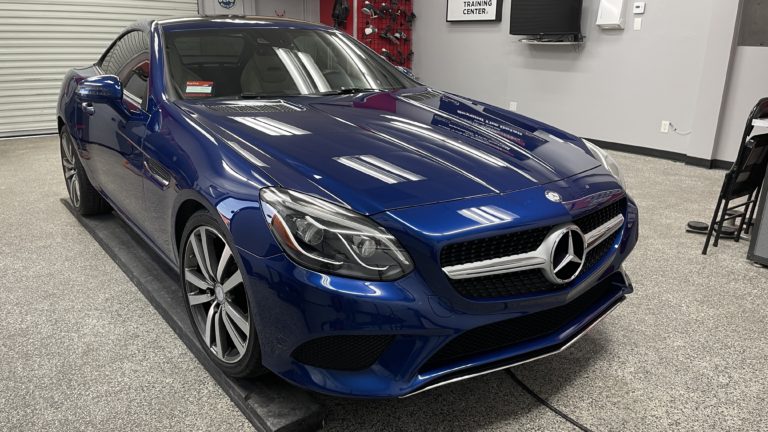 A sleek, blue Mercedes-Benz SL coupe is parked in a well-lit garage with a polished concrete floor. The car features a shiny exterior, prominent front grille with the Mercedes emblem, and stylish alloy wheels. Various equipment and a wall-mounted TV are visible in the background.