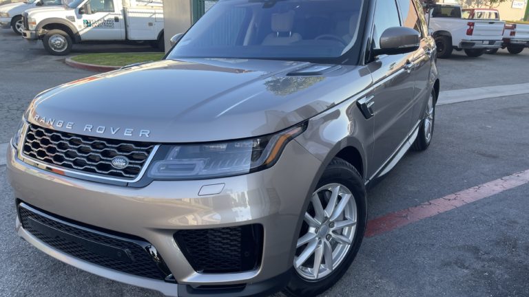 A silver Range Rover is parked on a street near a curb. The vehicle features a sleek, modern design with notable branding on the front grille. In the background, there are additional vehicles, including two white utility trucks, and a paved lot.