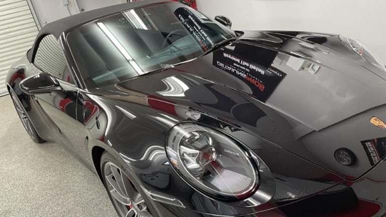 A black Porsche 911 convertible is parked in a garage. The car's sleek, polished body and distinctive headlights are prominently visible, reflecting the lighting from the garage. Various garage tools and equipment are in the background.