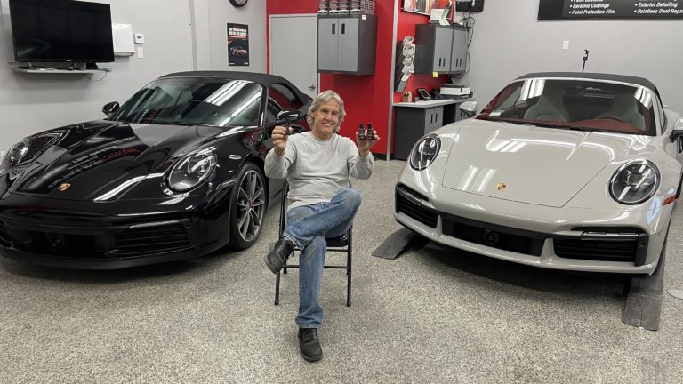 A person sitting on a chair in a garage, holding two objects in their hands, with a black sports car on the left and a silver sports car on the right. The garage has red and black cabinets and a workbench in the background. The person is smiling.