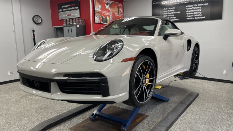 A sleek, silver sports car is elevated on a hydraulic lift in an auto detailing shop. The shop has gray floors, red and gray walls, and various detailing equipment and products. The car’s black and yellow brake calipers and large, shiny wheels are visible.