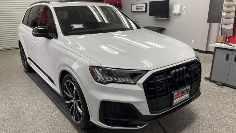 A white Audi SUV is parked in an indoor garage with a "Training Center" sign on the wall in the background. The vehicle has a sleek design, black window trim, and sporty rims. The garage floor has a speckled finish, and various equipment is visible in the background.