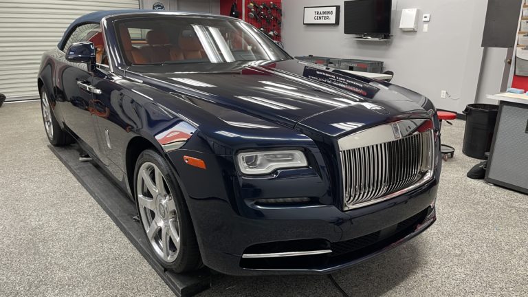 A sleek, dark blue Rolls-Royce convertible with a polished chrome grille and luxurious design is parked in a modern garage. The car's top is up, and the interior is partially visible. The garage features equipment, a "Training Center" sign, and a TV on the wall.