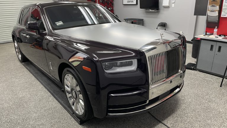 A black Rolls-Royce Phantom with a silver hood is parked in a garage on a gray floor. The car's sleek and shiny exterior is complemented by its iconic grille and Spirit of Ecstasy hood ornament. Tools and equipment are visible in the background.