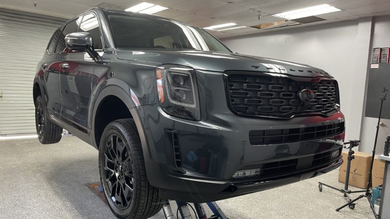 A dark gray SUV is elevated inside a garage or workshop. The vehicle's front grille and blacked-out rims are clearly visible. The workspace is equipped with various tools and equipment in the background, and a roller door is partially visible.