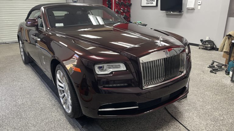 A dark maroon Rolls-Royce convertible is parked in a garage with a gray floor. The car has a shiny, polished exterior, a distinctive chrome grille with the Rolls-Royce emblem, and silver alloy wheels. The garage has various tools and equipment visible in the background.