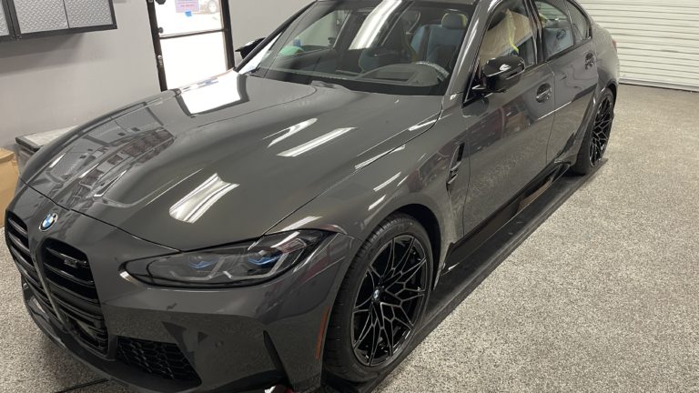 A sleek, dark gray BMW sedan is parked indoors on a smooth, grey-carpeted floor. The car features black alloy wheels, a modern design with sharp lines, and clear, reflective windows. The background shows a partially open roller door and various car-related equipment.