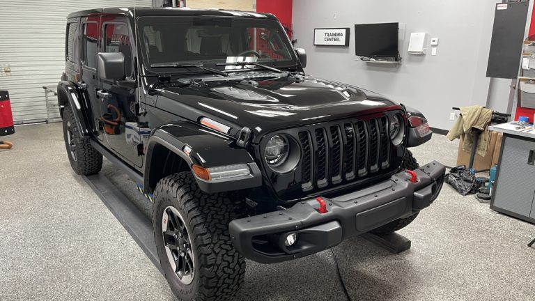 A black Jeep Wrangler is parked inside a garage on a lift. The vehicle has rugged tires, a distinctive seven-slot grille, and red tow hooks on the front bumper. The garage has a computer monitor and cabinets in the background, with a sign reading "Training Center.