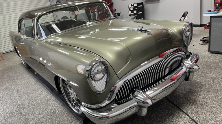 A vintage, olive green classic car is parked inside a garage. The vehicle has shiny chrome accents, a distinctive front grille, and rounded headlights. The garage setting includes a closed garage door, gray flooring, and assorted tools and equipment in the background.