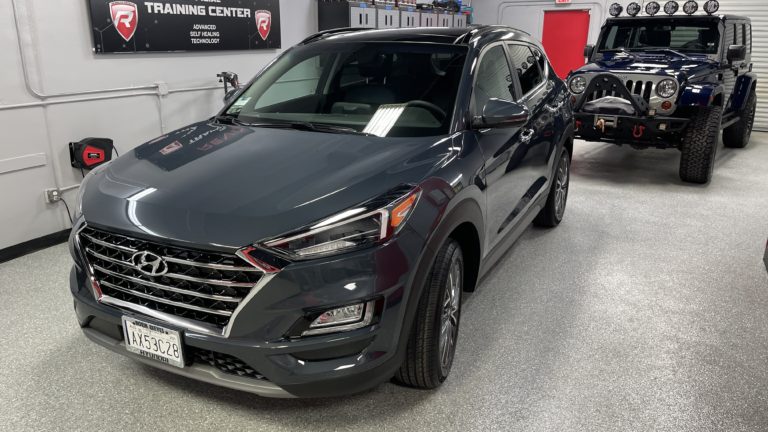 A gray Hyundai SUV is parked inside a garage with a textured floor. To the right, a lifted Jeep with large tires and additional lights on the roof is also visible. Various tools and equipment are mounted on the walls in the background.