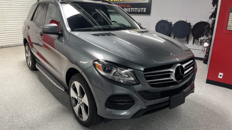 A gray Mercedes-Benz SUV parked indoors on a light-colored, speckled floor. The vehicle has a chrome grille, large alloy wheels, and a polished exterior. There is signage on the wall behind the car and cleaning equipment can be seen to the right.