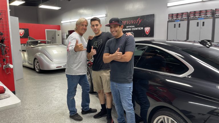 Three men are standing in a car detailing shop, giving thumbs-up gestures and smiling at the camera. Two black cars and garage equipment are visible in the background. One man is wearing a white long-sleeve shirt, another a black t-shirt, and the third a gray hat and black t-shirt.