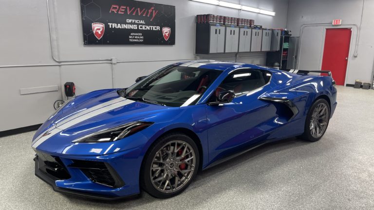 A shiny blue sports car is parked inside a modern garage with grey walls and cabinets. The car features sleek curves and a low profile. The walls display a "REVIV ify Official Training Center" sign and posters. A red door is visible in the background.