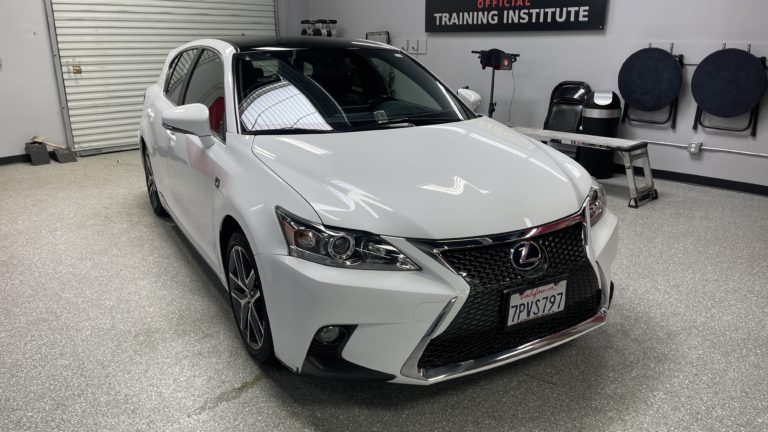 A white Lexus car is parked in a well-lit garage with a gray floor. The sign above the car reads "TRAINING INSTITUTE." The vehicle has sleek headlights, sporty design, and a visible license plate that reads "7PVS797." Car care supplies are visible in the background.