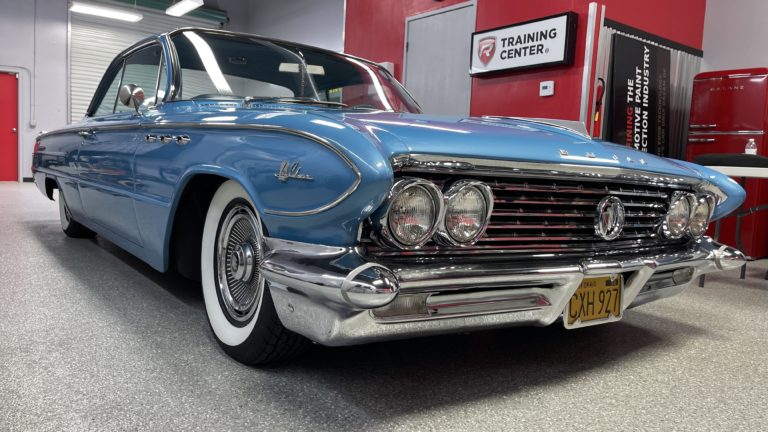 A beautifully maintained blue vintage car with chrome accents and whitewall tires is parked indoors. Behind it, a red and black training center is visible. The car's license plate reads "CMX927." The setting appears to be a classic car showroom or workshop.