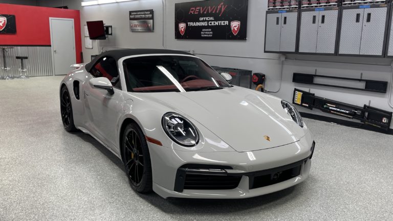 A sleek gray sports car with a black roof is parked in a pristine garage. The car has red interior accents, five-spoke black rims, and distinctively styled headlights. The garage features red and silver walls, organizational cabinets, and automotive equipment.