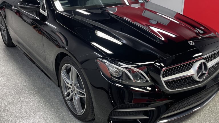 A sleek black Mercedes-Benz car, showcasing its front left side, headlight, and grille. The car is parked indoors with a grey speckled floor and white and red walls in the background. The vehicle's shiny surface reflects the room’s lighting and ambiance.