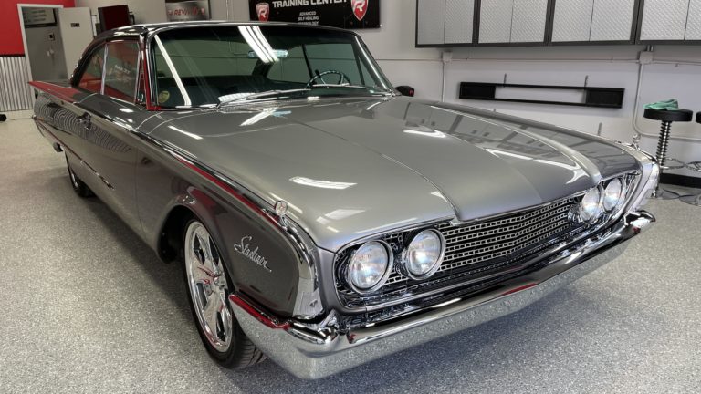 A sleek, vintage gray car with polished chrome detailing is parked inside a clean, well-lit garage. The car features a distinctive front grille with dual round headlights on each side and shiny, spoked wheels. The background includes tool cabinets and automotive equipment.