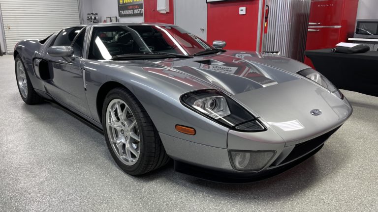 A sleek, silver Ford GT sports car is parked in a well-lit garage. The car has aerodynamic lines, a low profile, and large, detailed front headlights. A closed garage door and red walls with various automotive equipment are visible in the background.