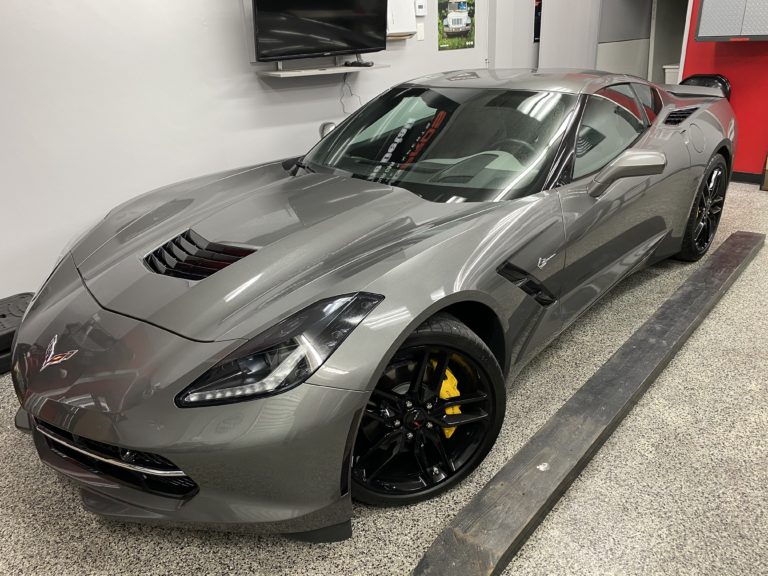 A sleek, metallic gray sports car with black rims and a streamlined design is parked indoors on a textured gray floor. The car features angular headlights, a vented hood, and a low profile. A wall-mounted TV and various equipment are visible in the background.