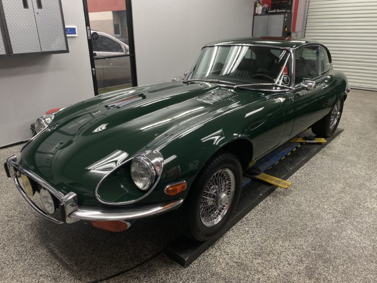 A classic green sports car, possibly a Jaguar E-Type, is parked in a well-lit garage. The car has a sleek design with noticeable chrome accents on the front bumper and wire-spoke wheels. It is positioned on a lift, and tools are visible in the background.