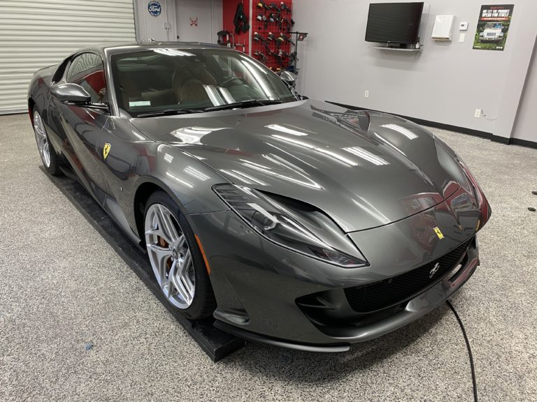 A sleek, dark gray Ferrari sports car is parked indoors on a polished concrete floor. The car features sharp, aerodynamic lines, silver alloy wheels, and a prominent Ferrari logo on the front grille. Background includes car care equipment and a wall-mounted TV.