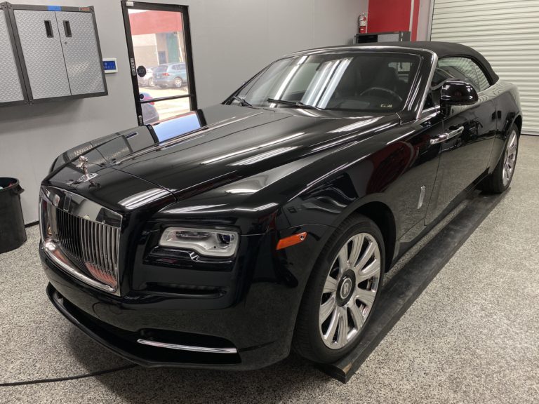 A sleek, black Rolls-Royce convertible is parked indoors on a slightly raised platform. The car has polished, reflective bodywork, silver trim, and distinctive Rolls-Royce grille. The background features grey walls, a roll-up door, and some industrial storage cabinets.