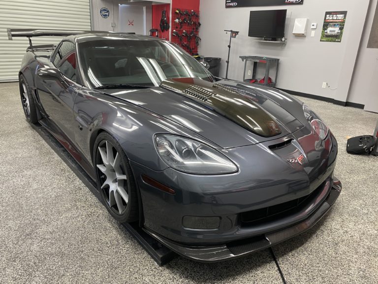 A sleek, dark gray sports car with a polished exterior is displayed indoors on a granite-like floor. The car features distinctive vents on the hood, a large rear wing, and appears to be in a professional garage setting with various tools and equipment in the background.