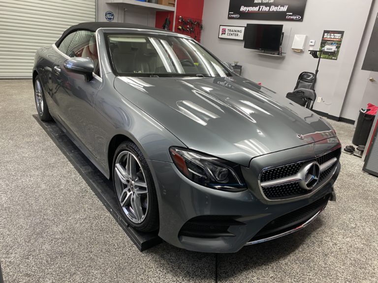 A sleek, silver Mercedes-Benz convertible is parked indoors, possibly in a garage or detailing shop. The car has a black soft top roof and polished bodywork. The setting includes automotive equipment and posters on the walls in the background.