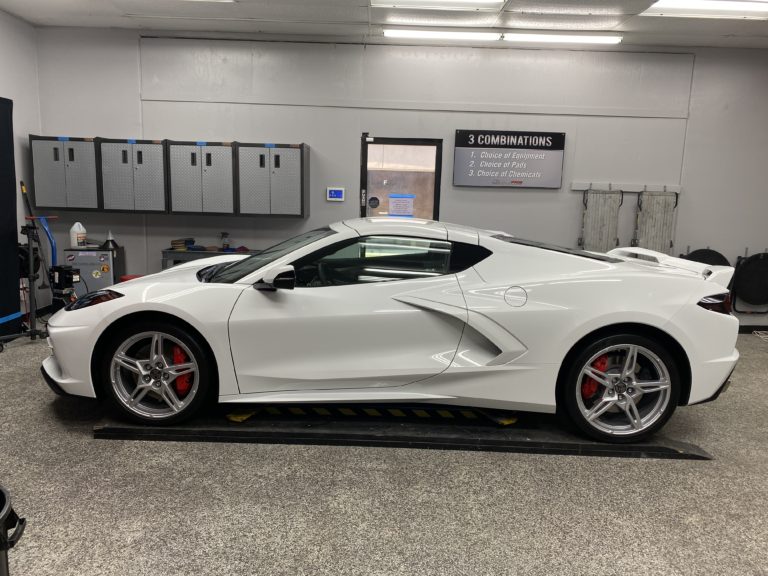 A white sports car with sleek, aerodynamic design is parked indoors on a slick shop floor. The car features red brake calipers, distinctive side air intakes, and silver alloy wheels. A sign in the background lists three combination options for choice and destinations.